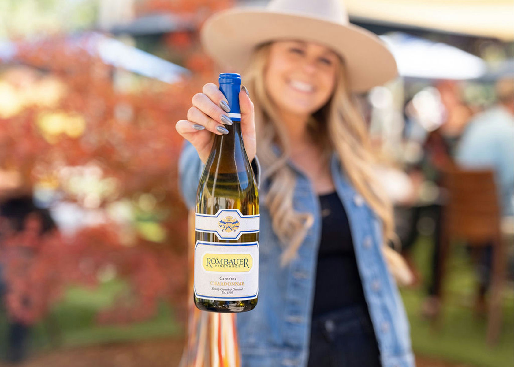Woman in cowboy hat holding up a bottle of Rombauer Chardonnay wine at an outdoor vineyard event.