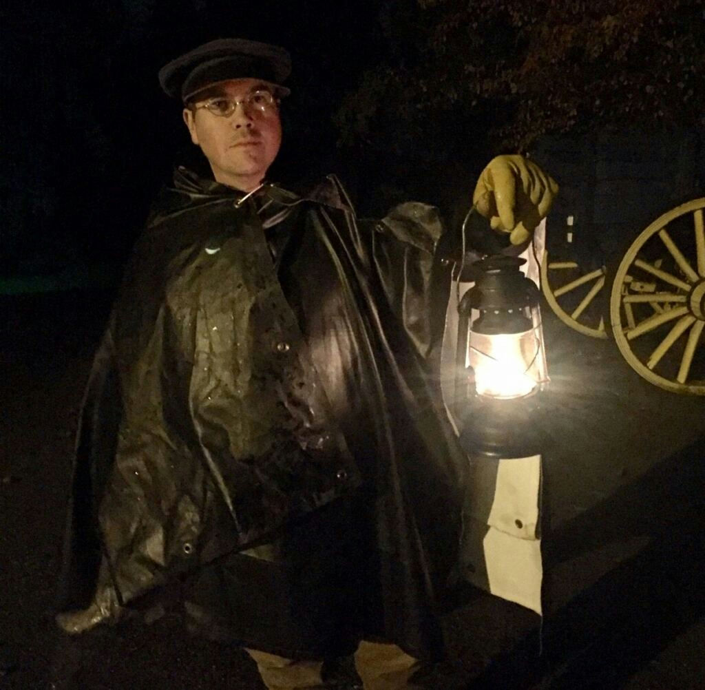 A person in period costume holds a glowing lantern at night beside a wagon wheel.